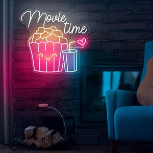 Op de afbeelding: Neon-bord met de tekst "Movie time" in wit cursief schrift. Het bord heeft ook een roze popcorn-emmer met een vrolijk gezicht en een blauwe beker met een rietje. Het bord is verlicht met roze en blauwe neonlichten.