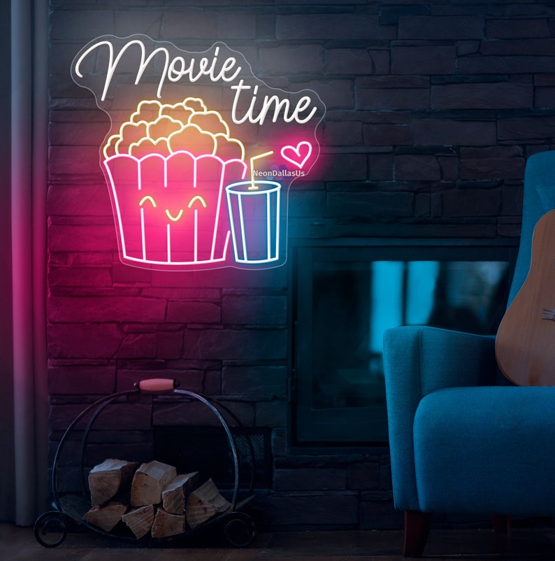 Op de afbeelding: Neon-bord met de tekst "Movie time" in wit cursief schrift. Het bord heeft ook een roze popcorn-emmer met een vrolijk gezicht en een blauwe beker met een rietje. Het bord is verlicht met roze en blauwe neonlichten.
