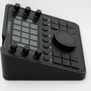 Könnte beinhalten: Schwarze Looqbok-Steuerungsoberfläche mit einem Raster aus Knöpfen, einem großen Drehknopf und einem Touchpad. Die Oberfläche hat eine schwarze Kunststoffbasis mit einer abgeschrägten Kante.