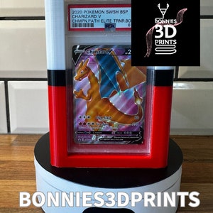 May include: A white, red and black 3D printed display case with a rotating base. The case holds a Pokemon trading card featuring Charizard V. The card is graded with a PSA 6. The display case has a label that reads "BONNIES 3D PRINTS".