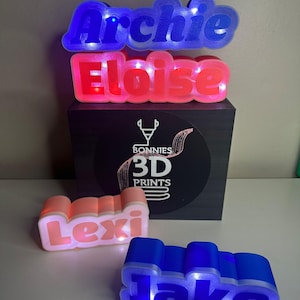 May include: Four personalized light-up name signs. The names are Archie, Eloise, Lexi, and Jake. The signs are made of 3D printed plastic and have a glossy finish. The lights are LED and change color. The signs are sitting on a black box with a white logo that says "Bonnies 3D Prints".