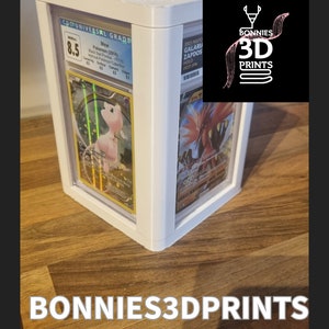 May include: White 3D printed display case for trading cards. The case has three slots for cards. The case is sitting on a wooden surface. The text "BONNIES3DPRINTS" is printed on the surface.