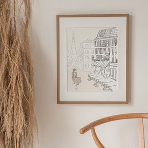 May include: A framed art print featuring a Parisian street scene with the Eiffel Tower in the background. The artwork is a line drawing with a woman in a dress riding a bicycle. The frame is made of light brown wood.