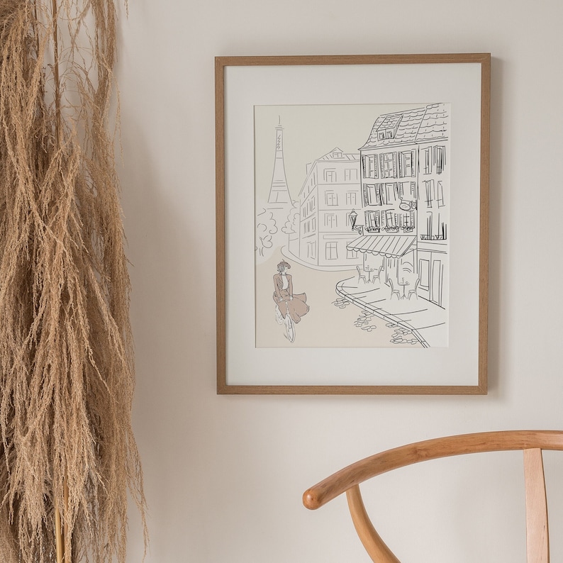 May include: A framed art print featuring a Parisian street scene with the Eiffel Tower in the background. The artwork is a line drawing with a woman in a dress riding a bicycle. The frame is made of light brown wood.