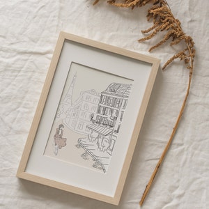 May include: Framed art print featuring a black and white line drawing of a Parisian street scene. The artwork includes the Eiffel Tower, buildings, and a person riding a bicycle. The frame is light wood, and the print has a neutral background.