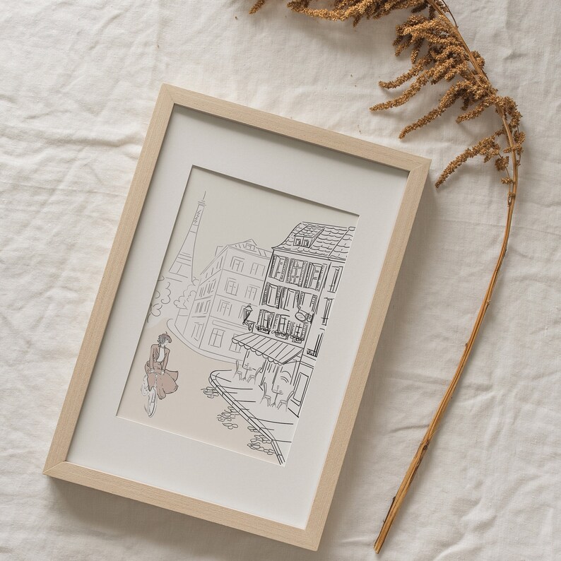 May include: Framed art print featuring a black and white line drawing of a Parisian street scene. The artwork includes the Eiffel Tower, buildings, and a person riding a bicycle. The frame is light wood, and the print has a neutral background.