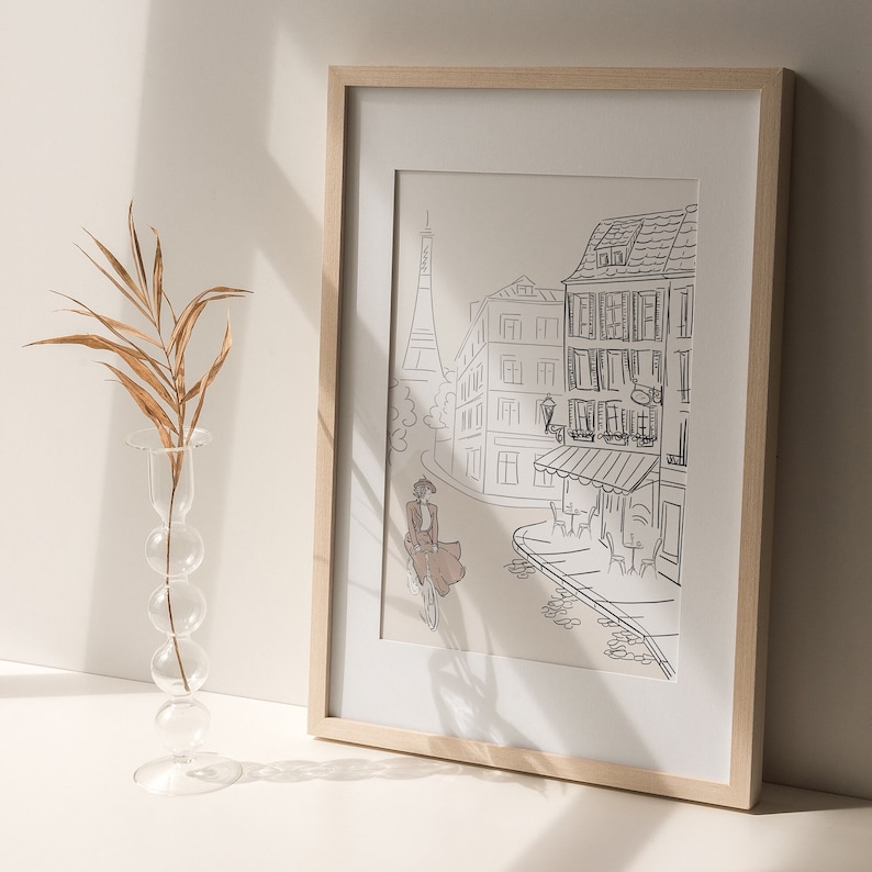 May include: A framed print featuring a black and white line drawing of a Parisian street scene. The artwork depicts the Eiffel Tower, buildings, and a person riding a bicycle. The frame is light-colored wood, and the print is matted.