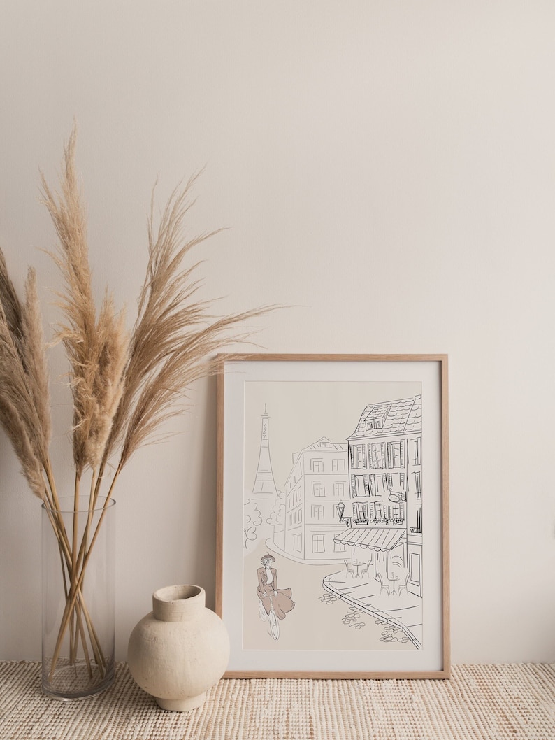 May include: A framed art print featuring a line drawing of a Parisian street scene with the Eiffel Tower in the background. The artwork depicts a person riding a bicycle, with buildings and a cafe. The frame is made of light-colored wood.