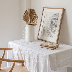 May include: A framed black and white print of a Parisian street scene, a white vase with a dried palm leaf, a glass candle holder, and two books with a gold-colored bar on a white tablecloth. A wooden chair is in the foreground.