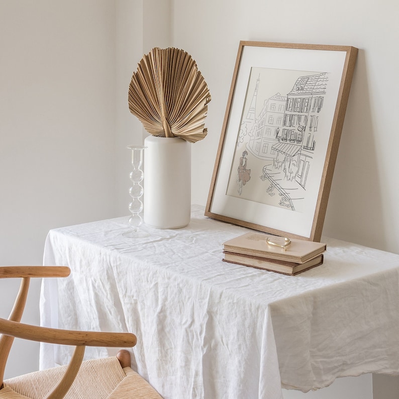 May include: A framed black and white print of a Parisian street scene, a white vase with a dried palm leaf, a glass candle holder, and two books with a gold-colored bar on a white tablecloth. A wooden chair is in the foreground.