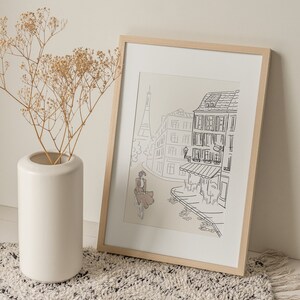 May include: A framed art print featuring a black and white line drawing of a Parisian street scene with the Eiffel Tower. The print is in a light wood frame. A white vase with dried flowers sits to the left of the print.