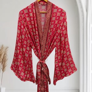 May include: A red patterned robe with long sleeves hangs on a wooden hanger. The robe features a repeating geometric design in shades of red and gold. The robe is tied at the waist with a matching fabric belt.