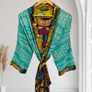 Może przedstawiać: Kolorowy, wzorzysty szlafrok z wiązaniem. Szlafrok ma turkusowe tło z geometrycznym wzorem i żywe wnętrze z żółtymi, fioletowymi i czarnymi wzorami. Szlafrok wisi na drewnianym wieszaku.