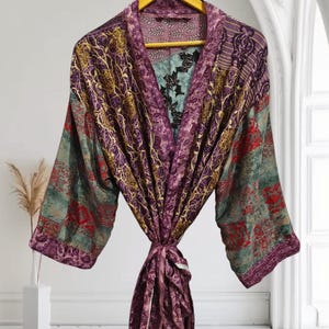 Op de afbeelding: Een kleurrijke, patroonrijke kimono met een striksluiting. De kimono heeft een mix van paarse, groene en gouden tinten met bloemen- en paisley-ontwerpen. Het hangt aan een gele hanger tegen een witte achtergrond.