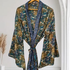 May include: A lightweight kimono robe featuring a detailed paisley pattern in shades of teal, gold, and cream. The garment has wide, flowing sleeves and a contrasting dark blue border along the front opening and hem, tied with a matching fabric belt.