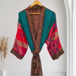 May include: A long-sleeved kimono robe featuring a patchwork design with vibrant red, teal, and dark brown sections. The garment displays a mix of intricate floral and paisley patterns, a wide collar, and a matching tie belt, all hanging on a wooden hanger.