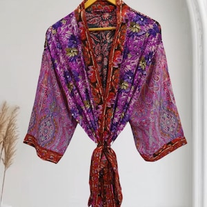 May include: A vibrant, floral-patterned robe in shades of purple, pink, and red hangs on a wooden hanger. The robe features a paisley design on the sleeves and a contrasting floral pattern on the inner lining. The robe is tied at the waist with a matching fabric belt.