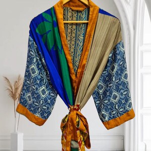 Può includere: Una colorata vestaglia kimono con un design patchwork. La vestaglia presenta un mix di tessuti blu, verdi, beige e arancioni con motivi floreali e geometrici. È appesa a una gruccia di legno su uno sfondo bianco.
