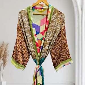 May include: A kimono-style robe featuring a patchwork design with contrasting panels of gold floral patterns, geometric shapes in vibrant colors like pink, blue, and orange, and solid green trim, hanging on a wooden hanger with long sleeves.