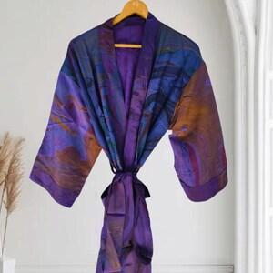 Könnte beinhalten: Ein langärmeliger Morgenmantel mit einem lebendigen Marmormuster in tiefen Violett-, Blau- und Orangetönen. Das Kleidungsstück hat einen breiten Kragen, einen passenden Bindegürtel in der Taille und eine glatte, satinartige Oberfläche am Kleiderbügel.