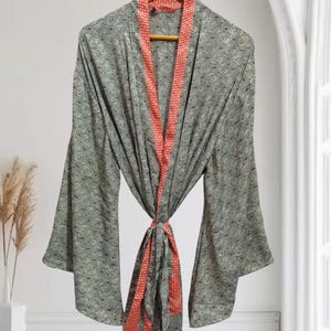 May include: A gray patterned robe with wide sleeves and a coral-colored trim and tie. The robe is hanging on a wooden hanger against a white background. The robe's pattern is a repeating geometric design.