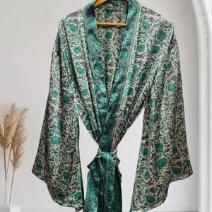 May include: A long-sleeved robe featuring a repeating floral pattern in shades of emerald green and dark grey on a light beige background. The garment includes a wide shawl collar, a matching tie belt at the waist, and wide, flowing sleeves.