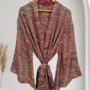 May include: A long-sleeved robe featuring a deep red base colour with an intricate, multi-coloured paisley pattern throughout. The garment includes a matching fabric tie at the waist and wide, flowing sleeves, all displayed on a wooden hanger.