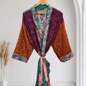 Może przedstawiać: Szlafrok typu kimono z długimi rękawami, wykonany w stylu patchworkowym w żywych kolorach, takich jak pomarańczowy, fioletowy, różowy i turkusowy. Odzież posiada skomplikowane wzory kwiatowe i paisley oraz pasujący pasek w talii.