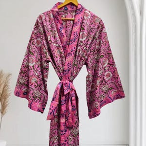 Könnte beinhalten: Ein leuchtend pinkfarbener Morgenmantel im Kimono-Stil mit einem detaillierten Blumen- und Blattmuster in Weiß, Dunkelblau und Grün. Das Kleidungsstück hat weite Ärmel, einen passenden Bindegürtel und einen Schalkragen und hängt an einem Holzkleiderbügel.