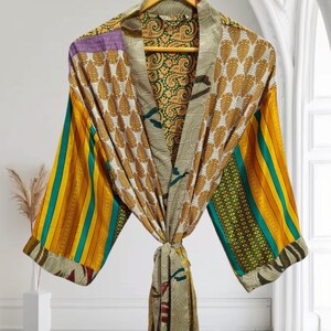 May include: A colorful, patterned robe hanging on a wooden hanger. The robe features a mix of patterns, including leaf and paisley designs, and stripes in shades of yellow, green, and purple. The robe is tied at the waist.