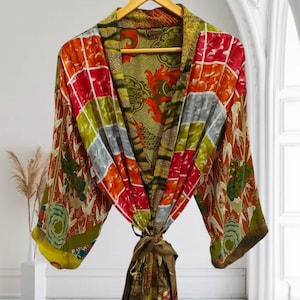 Könnte beinhalten: Ein farbenfroher, gemusterter Bademantel mit Bindverschluss, der an einem Holzbügel hängt. Der Bademantel hat ein Patchwork-Design mit roten, orangefarbenen, grünen und grauen Quadraten. Das Innenfutter hat ein Blumenmuster. Die Ärmel haben ein Blumen- und Blätterdesign.