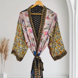 Puede incluir: Una bata tipo kimono de manga larga con un diseño de patchwork que presenta un panel central floral rosa claro, mangas con flores amarillas y verdes, y un borde negro con patrones geométricos dorados. Incluye un cinturón a juego en la cintura.