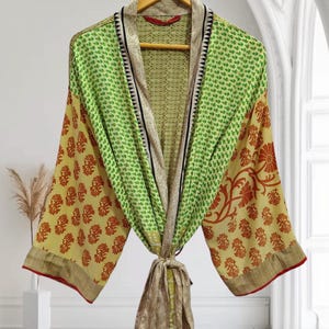 May include: A colorful kimono robe with a floral pattern. The robe features a mix of green, yellow, and red hues, with a tie closure at the waist. The robe is hanging on a wooden hanger.