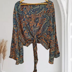 May include: A long-sleeved top with a paisley pattern in shades of orange, blue, and green. The top features a square neckline and a tie detail at the waist. The sleeves are flared, and the top is hanging on a wooden hanger.
