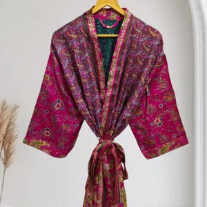 Può includere: Un'accappatoio rosa acceso con motivo paisley e interno verde, appeso a una gruccia di legno. L'accappatoio presenta una chiusura con fiocco ed è realizzato in un tessuto setoso. Il design include motivi floreali e paisley.