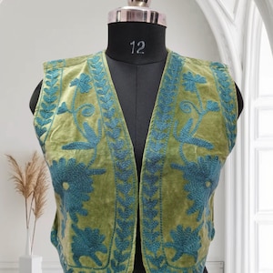 Peut inclure: Un gilet en velours vert avec des broderies bleues de motifs floraux. Le gilet a un col en V et est sans manches.