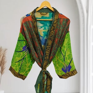 May include: A colorful, patterned robe with a tie closure, hanging on a wooden hanger. The robe features a mix of green, orange, and blue fabrics with paisley and floral designs. The sleeves are long and the robe appears to be made of a lightweight material.