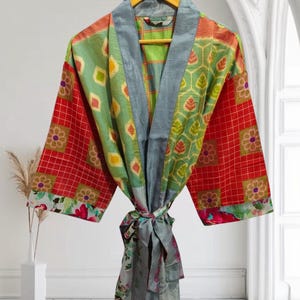 May include: A colorful patchwork robe with a tie closure. The robe features a mix of patterns, including red squares with floral accents, green diamond shapes, and floral and leaf designs. The robe is hanging on a wooden hanger.