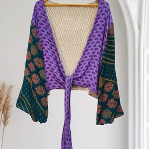 May include: A long-sleeved wrap top featuring a mix of fabrics. The centre panels are purple with a small repeating pattern, while the sleeves are dark green with a large orange leaf print. A beige chevron-patterned fabric lines the back.