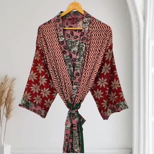 Könnte beinhalten: Ein langärmeliger Kimono-Bademantel mit Patchwork-Design in tiefroten, rosafarbenen und dunkelgrünen Tönen. Der Stoff zeigt ein wiederkehrendes Muster aus hellen sternförmigen Blättern und Zickzackstreifen, ergänzt durch einen passenden Bindegürtel.