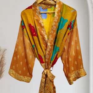 May include: A vibrant, flowing robe in shades of gold, orange, and brown, adorned with floral patterns in red, blue, and green. The robe features wide sleeves and a tie closure at the waist. The garment is hanging on a wooden hanger.