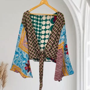 May include: A cropped wrap top with long bell sleeves, featuring a patchwork design of various patterns including green polka dots, blue floral motifs, and red accents. The garment is tied at the front with a long fabric sash and hangs on a hanger.
