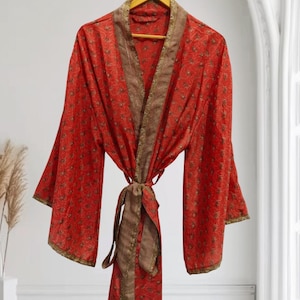Może przedstawiać: Czerwone szlafrok w stylu kimono z kwiatowym wzorem i brązowymi wykończeniami, wiszący na drewnianym wieszaku. Szlafrok jest zawiązany w talii pasującym paskiem. Rękawy są szerokie i zwiewne, a materiał wydaje się lekki.