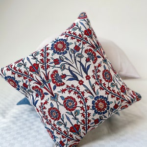 May include: A white throw pillow with a red, blue, and green floral pattern. The pillow is made of a soft, woven fabric and has a square shape.