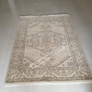 Peut inclure: Un tapis rectangulaire beige avec un motif g&eacute;om&eacute;trique d&eacute;taill&eacute; dans des tons bruns. Le tapis pr&eacute;sente un motif central avec une bordure et le texte "NAZAR CARPETS". Le tapis est pos&eacute; sur un sol clair.