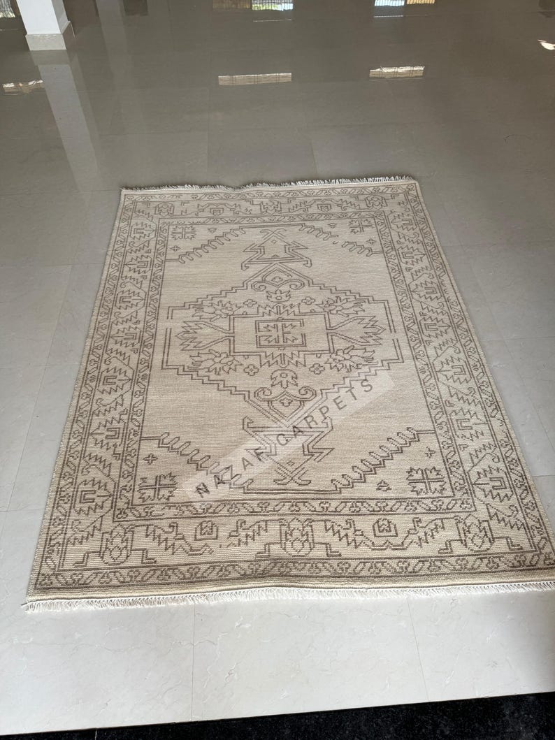Peut inclure: Un tapis rectangulaire beige avec un motif g&eacute;om&eacute;trique d&eacute;taill&eacute; dans des tons bruns. Le tapis pr&eacute;sente un motif central avec une bordure et le texte "NAZAR CARPETS". Le tapis est pos&eacute; sur un sol clair.