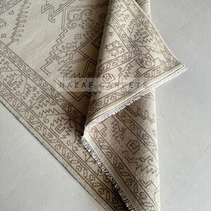 Peut inclure: Tapis beige et marron &agrave; motifs g&eacute;om&eacute;triques. Le tapis est partiellement enroul&eacute;, r&eacute;v&eacute;lant les d&eacute;tails complexes du motif. Le texte "NAZAR CARPETS" est visible sur le tapis.