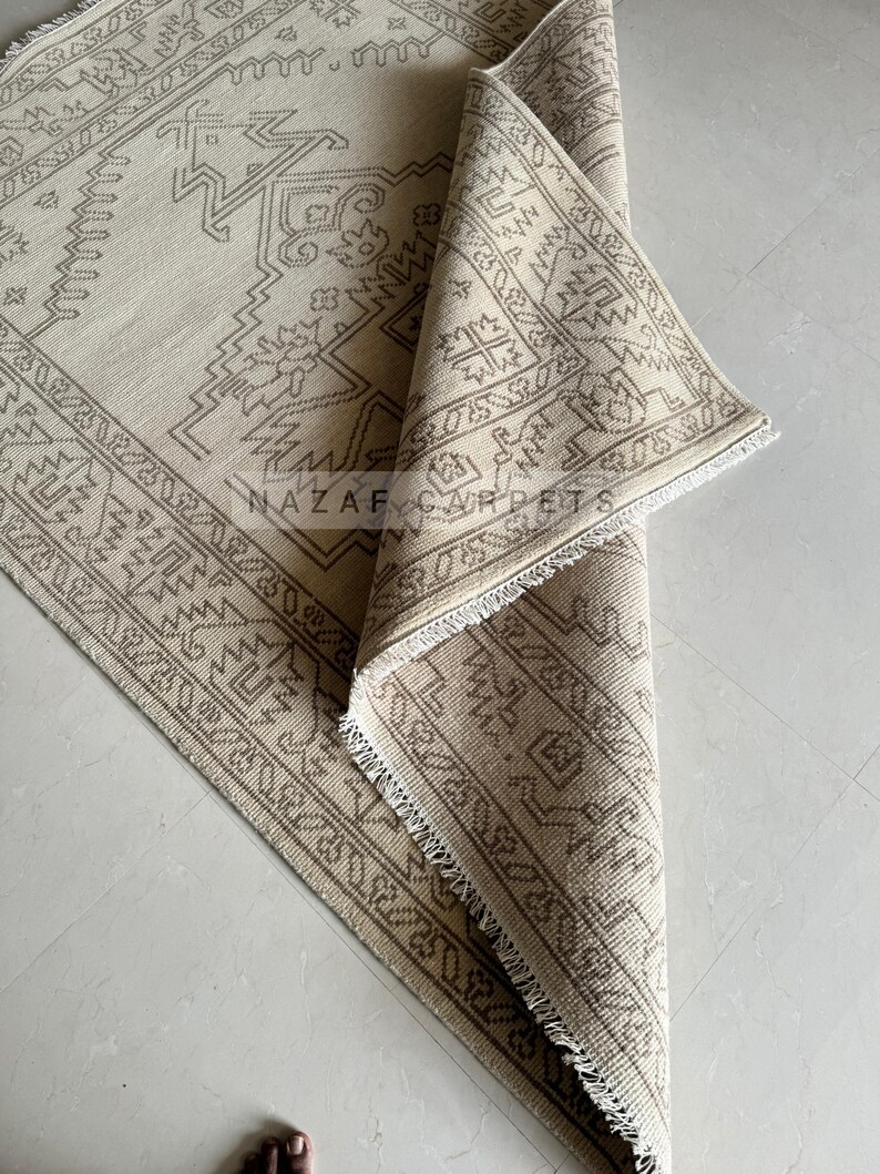 Peut inclure: Tapis beige et marron &agrave; motifs g&eacute;om&eacute;triques. Le tapis est partiellement enroul&eacute;, r&eacute;v&eacute;lant les d&eacute;tails complexes du motif. Le texte "NAZAR CARPETS" est visible sur le tapis.