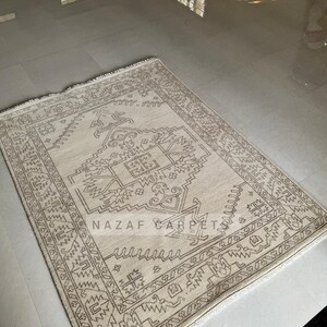 Peut inclure: Tapis rectangulaire beige avec un motif g&eacute;om&eacute;trique dans les tons marron. Le tapis pr&eacute;sente un motif central et une bordure d&eacute;taill&eacute;e. Le texte "NAZAF CARPET" est visible sur le tapis.
