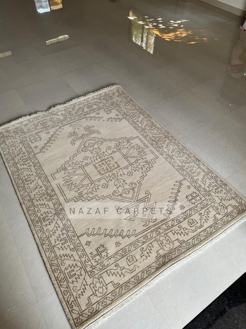 Peut inclure: Tapis rectangulaire beige avec un motif g&eacute;om&eacute;trique dans les tons marron. Le tapis pr&eacute;sente un motif central et une bordure d&eacute;taill&eacute;e. Le texte "NAZAF CARPET" est visible sur le tapis.